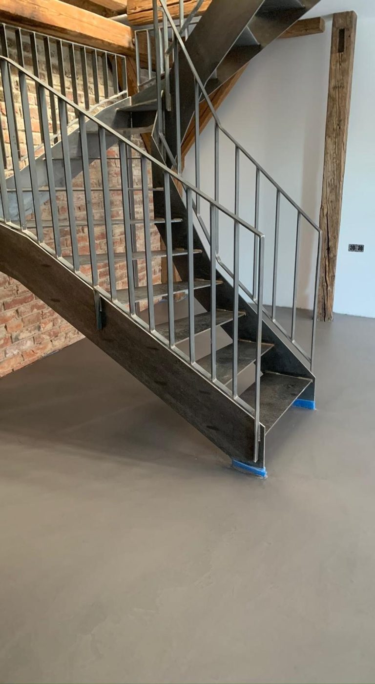 Treppenhaus mit schwarzer Treppe und metallischem Geländer, auf einem grauen Mikrozement Boden.